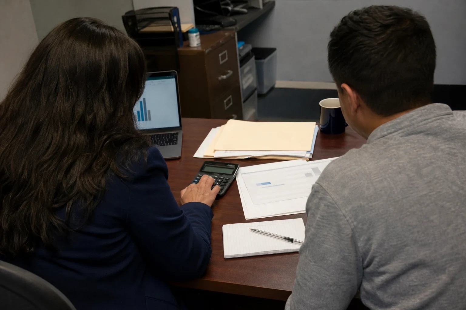 Over-the-shoulder view of two people at an office desk reviewing printed statements, one using a calculator beside a notepad, folders, laptop and filing cabinet