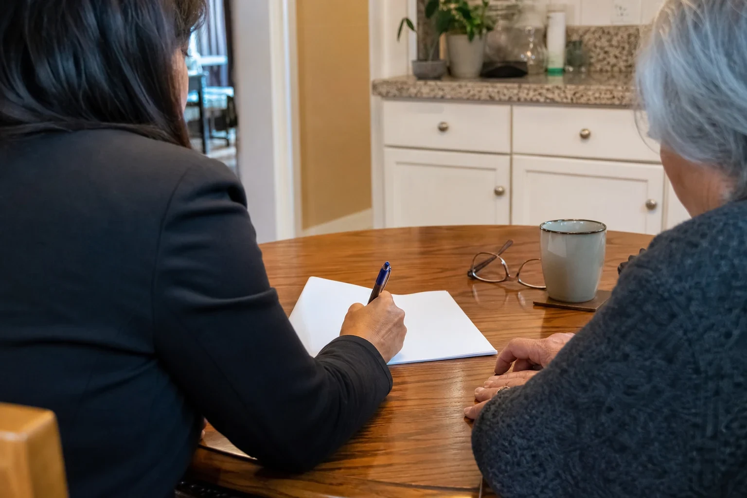 Agent in a dark blazer writes on a blank form at a wooden kitchen table beside an older gray-haired woman, with reading glasses and a mug nearby