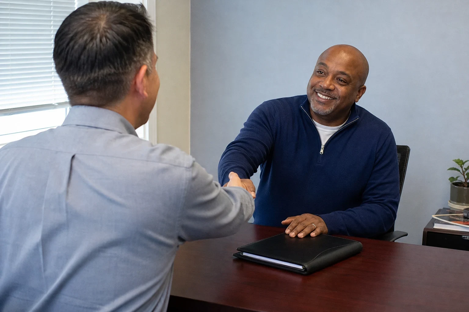 Insurance agent in a light blue shirt shakes hands across a wooden desk with a smiling client in a navy sweater, a closed black folder on the desk