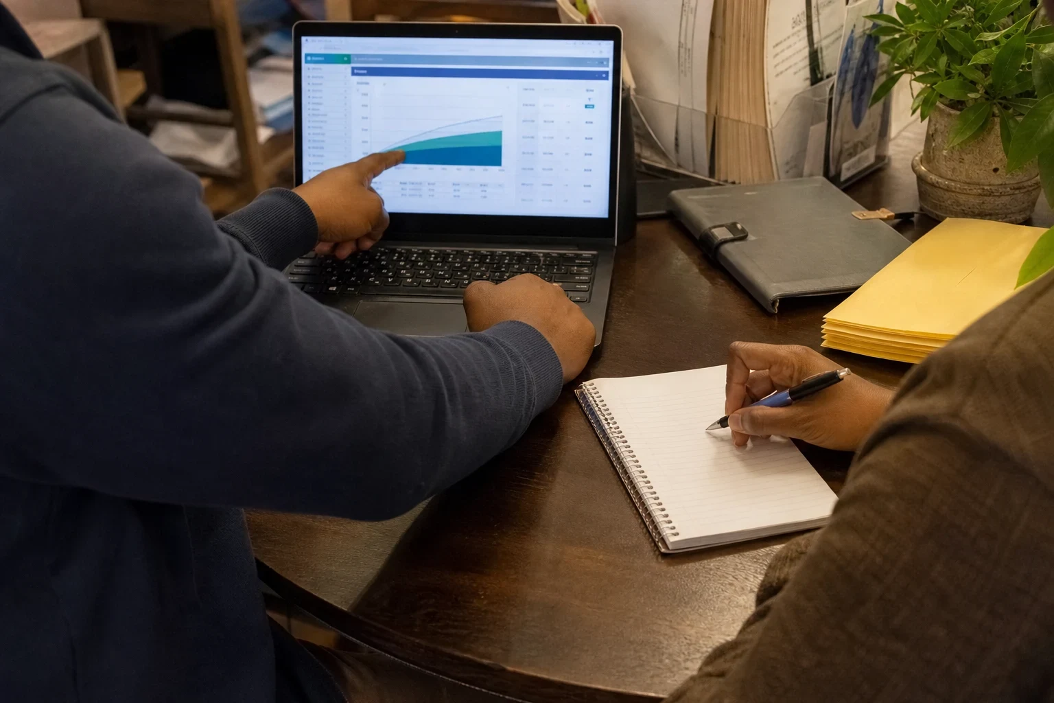 An agent points to a growth chart on a laptop at a wooden office desk while a client takes notes in a spiral notebook beside yellow envelopes and a plant.