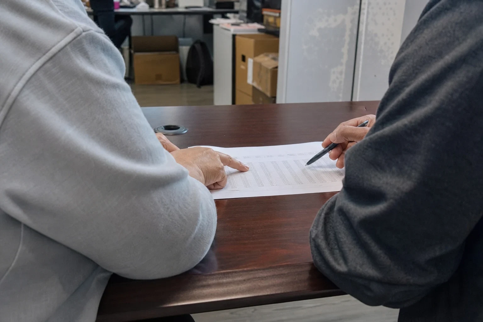 Two people at a dark wood conference table, one pointing at a printed table of figures while the other holds a pen, with boxes and office shelving behind them